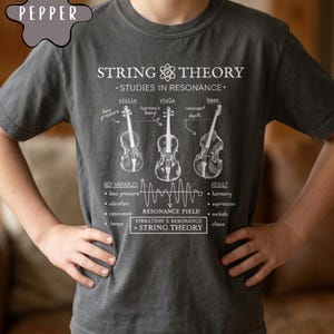 May include: A charcoal gray t-shirt featuring a white graphic print titled String Theory Studies in Resonance. The design includes illustrations of a violin, viola, and bass, along with text labels, a wave diagram, and the equation Vibration + Resonance = String Theory.