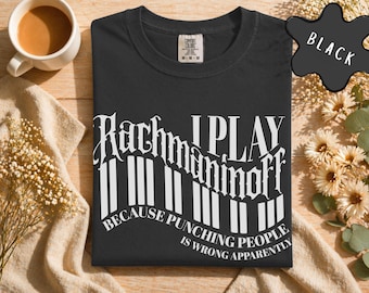 May include: Black t-shirt with white text that reads "Rachmaninoff I PLAY because punching people is wrong apparently." The text is designed to look like piano keys. The shirt is folded on a wooden surface.