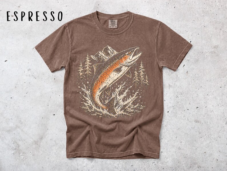K&ouml;nnte beinhalten: Ein espressofarbenes Kurzarm-T-Shirt mit einem Grafikdruck eines aus dem Wasser springenden Fisches. Das Design zeigt im Hintergrund ein Gebirge und Tannenb&auml;ume in wei&szlig;en und orangefarbenen T&ouml;nen. Der Text ESPRESSO ist auf dem Bild zu sehen.