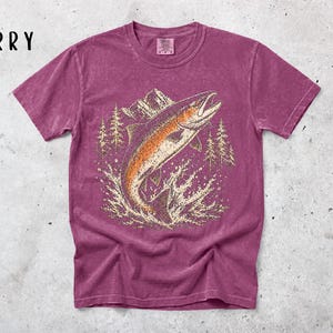 K&ouml;nnte beinhalten: Ein beerenfarbenes Kurzarm-T-Shirt mit einem Grafikdruck eines springenden Fisches im Wasser, umgeben von Tannen und Bergen. Das Wort BERRY ist in schwarzer Schrift in der oberen linken Ecke des Bildbereichs aufgedruckt.