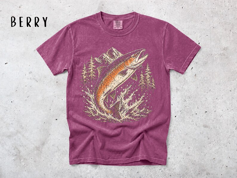 K&ouml;nnte beinhalten: Ein beerenfarbenes Kurzarm-T-Shirt mit einem Grafikdruck eines springenden Fisches im Wasser, umgeben von Tannen und Bergen. Das Wort BERRY ist in schwarzer Schrift in der oberen linken Ecke des Bildbereichs aufgedruckt.