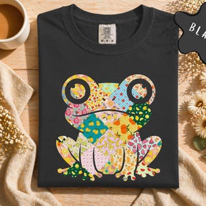 May include: Black t-shirt featuring a colorful patchwork frog design. The frog's body is composed of various patterned patches with fruits and floral motifs. The shirt has a classic crew neck and short sleeves. The tag reads "Comfort Colors".