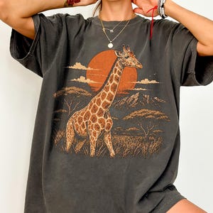 May include: A charcoal gray short-sleeve t-shirt featuring a graphic print of a giraffe standing in a savanna landscape with a large orange sun, mountains, and trees in the background. The word PEPPER is printed in the upper right corner.