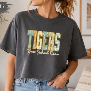 May include: A charcoal grey short-sleeve t-shirt featuring the word TIGERS in large block letters with varied patterns including stripes, floral, and gingham prints. Below the word TIGERS, the text Your School Name is printed in a cursive font.