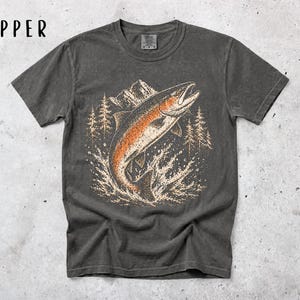 K&ouml;nnte beinhalten: Ein anthrazitfarbenes Kurzarm-T-Shirt mit einem Grafikdruck eines springenden Fisches in Orange- und Cremet&ouml;nen vor einer Kulisse aus Bergen und Tannen. Das Wort PEPPER ist in Schwarz in der oberen linken Ecke des Bildes aufgedruckt.