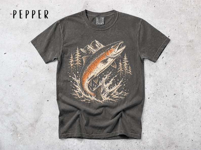 K&ouml;nnte beinhalten: Ein anthrazitfarbenes Kurzarm-T-Shirt mit einem Grafikdruck eines springenden Fisches in Orange- und Cremet&ouml;nen vor einer Kulisse aus Bergen und Tannen. Das Wort PEPPER ist in Schwarz in der oberen linken Ecke des Bildes aufgedruckt.
