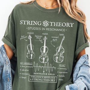 May include: An olive green t-shirt featuring a white graphic print titled STRING THEORY. The design includes illustrations of a violin, viola, and bass with labels like bow pressure and resonant depth, plus a resonance field wave and text equations.