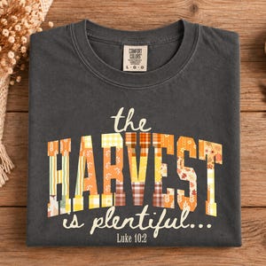 Peut inclure: Un t-shirt à col rond gris anthracite avec le texte "the HARVEST is plentiful... Luke 10:2" en police couleur crème. Le mot "HARVEST" est rempli d'un motif patchwork composé de carreaux orange, jaunes et blancs et de motifs de feuilles d'automne.