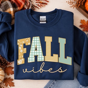 Puede incluir: Una sudadera azul marino de cuello redondo con el texto FALL en letras grandes con patrones variados que incluyen flores, cuadros vichy y rayas, y la palabra vibes en letra cursiva debajo, impresa en una prenda de la marca Gildan.
