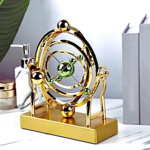 Könnte beinhalten: Eine goldfarbene, kinetische Skulptur mit kreisförmigem Design. Die Skulptur hat mehrere konzentrische Ringe und Kugeln in Gold, mit grünen Kugeln in der Mitte. Sie steht auf einem rechteckigen Sockel.