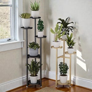 Op de afbeelding: Twee plantenstandaards met meerdere niveaus. De ene is zwart en de andere goudkleurig. Elke standaard bevat verschillende potplanten met groen blad. De standaards staan in een hoek bij een raam, met een houten vloer.