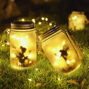 Può includere: Due barattoli di vetro Mason con sagome di fate e luci a stringa all'interno, su erba verde. I barattoli hanno coperchi in metallo e manici in corda. Luci aggiuntive sullo sfondo creano un'atmosfera magica.