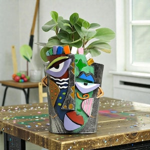 May include: A decorative face-shaped planter with a colourful, abstract design. The planter features a variety of colours, including purple, green, blue, and orange. It holds a green leafy plant. The planter is on a wooden table.