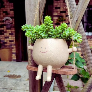 Puede incluir: Una maceta de cerámica beige caprichosa con forma de personaje sonriente, con brazos, piernas y un columpio. La maceta está llena de suculentas verdes vibrantes. El columpio es de madera y está suspendido por una cuerda.