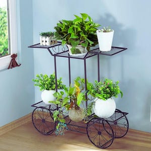 Puede incluir: Un soporte para plantas de metal negro con tres niveles, cada uno con plantas en macetas con follaje verde. El soporte tiene una base decorativa con ruedas y está colocado contra una pared azul claro. Las plantas están en macetas blancas.
