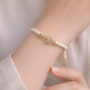 May include: Cream-colored rope bracelet with a gold-colored metal clasp and a small letter "K" charm encrusted with clear crystals. The bracelet is on a wrist.