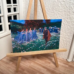 Può includere: Un dipinto su un cavalletto in miniatura raffigura un gruppo di donne in abiti blu fluenti che ballano in un campo di fiori bianchi. Un uomo in un cappotto marrone si trova al centro del dipinto, osservando le donne.