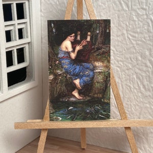 May include: A miniature painting on a wooden easel depicts a woman in a blue gown playing a lyre. The artwork has a classical style, set in a natural, wooded environment with a water feature.