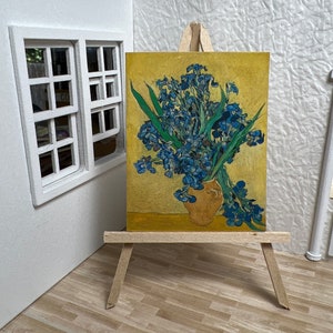 Puede incluir: Una pintura de un ramo de iris azules en un jarrón, sobre un fondo amarillo. La pintura se muestra en un caballete de madera.