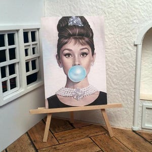 May include: A miniature painting of a woman blowing a bubble gum bubble, displayed on a small wooden easel. The woman has dark hair, a pearl necklace, and is wearing a black dress. The background is a light color.