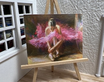 May include: A painting on a wooden easel depicts a ballerina in a pink tutu, tying her ballet shoes. The ballerina is posed in a seated position with her legs crossed. The painting is set against a soft, blurred background.