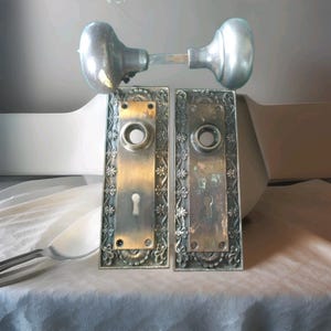 May include: A pair of rectangular brass door plates with ornate floral borders, each featuring a central circular knob hole and a keyhole. Above the plates sits a matching set of two round, metallic door knobs connected by a square spindle.
