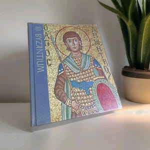 May include: A hardcover book titled Byzantium from the Great Ages of Man series. The cover features a gold mosaic illustration of a figure in armor holding a spear and shield, with Greek text flanking the figure on a light blue spine.