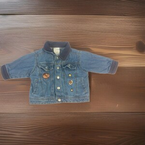 May include: A blue denim jacket featuring a dark navy collar and sleeve cuffs, silver-toned metal buttons, two chest pockets, and small decorative patches including a brown oval, a football, and two gold-colored stars on the front panel.