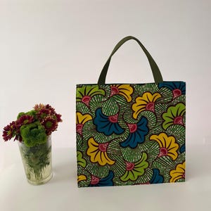 Pode incluir: Uma sacola quadrada com uma alça verde e um padrão floral vibrante em amarelo, azul e verde. O design da sacola apresenta um padrão repetido de flores estilizadas em um fundo verde. A sacola é feita de tecido.