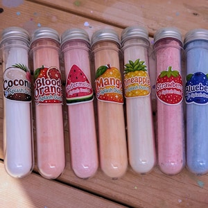 May include: Seven clear plastic tubes with silver lids, each filled with colourful powder. The tubes are labelled with fruit names like Coconut, Blood Orange, Watermelon, Mango, Pineapple, Strawberry, and Blueberry. The tubes are arranged on a wooden surface.