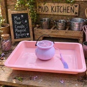 May include: A pink rectangular tray holds a small, light purple cauldron with a black handle and a purple and blue tassel. A chalkboard with text that reads "Mix, Pour, Create, Magical Recipes" is in the background. The scene is set on a wooden surface with other play kitchen accessories.