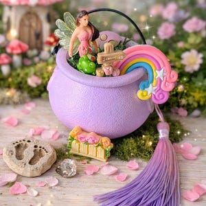 Puede incluir: Una escena de jardín de hadas con un caldero morado, un arcoíris, una hada y figuras de rana. Un letrero dice "Flowers". Una borla cuelga del caldero. Otros elementos incluyen una casa de champiñones, una piedra y pétalos de flores.