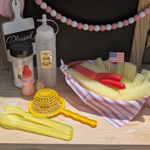 May include: A toy kitchen set featuring a red and white striped basket filled with yellow foam fries and red plastic hot dogs, a small American flag, a yellow plastic strainer, two yellow spoons, a clear squeeze bottle labeled Magic Oil, and a shaker.