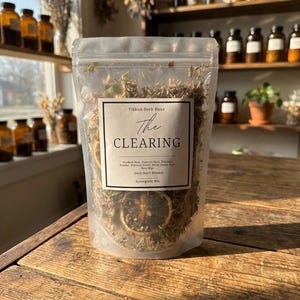 May include: A clear, resealable bag filled with dried herbs and citrus slices. The label reads "Tikkun Herb Haus The CLEARING" with a list of ingredients. The bag sits on a wooden surface, with jars and shelves in the background.