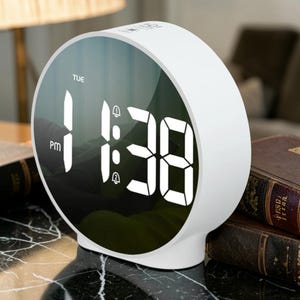 May include: A white, round digital alarm clock displaying the time as 1:38 PM. The clock face is a reflective black surface with white numerals and the day of the week, Tuesday, displayed at the top. Two bell icons are also visible.