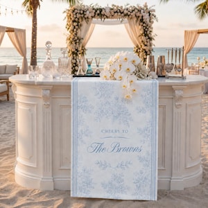 May include: A white bar with a marble top on a beach, decorated for a wedding. A floral runner with blue floral patterns and the text "Cheers to The Browns" is on the bar. The bar has glassware, a bouquet of white orchids, and bar tools.