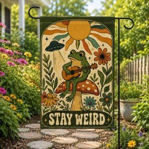 Könnte beinhalten: Eine rechteckige Gartenflagge mit einem grünen Frosch, der auf einem gepunkteten Pilz sitzt und Gitarre spielt. Über dem Frosch befinden sich eine fliegende Untertasse und eine Sonne. Das Design zeigt Blumen und den Text STAY WEIRD in Retro-Schrift.