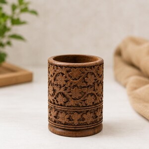 May include: A cylindrical wooden container with intricate floral carvings. The dark brown piece features detailed relief patterns of flowers and vines, with a smooth, unadorned interior. The container sits on a small, circular base.