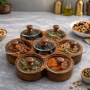 May include: A flower-shaped wooden serving tray featuring seven circular compartments, each topped with a clear glass lid and a small brown wooden knob. The compartments are filled with various nuts, dried fruits, and seeds, arranged in a cluster.