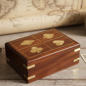 May include: A rectangular wooden playing card box with brass detailing and suit symbols inlaid on the lid. The box is on a light-coloured wooden surface. A red velvet bag and playing cards are also visible.