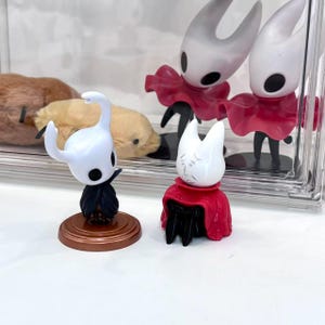 May include: A collection of small figurines featuring characters with white heads, black capes, and red cloaks. One figurine stands on a brown base. The figures are displayed in front of a clear, rectangular container.