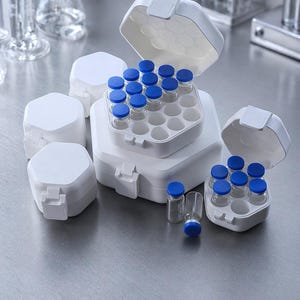 May include: A collection of white hexagonal storage cases for small glass vials. The cases feature hinged lids and interior compartments designed to hold vials securely. Several vials with blue caps are visible, both inside the open cases and standing upright on the surface.