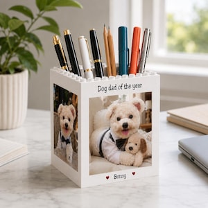 Puede incluir: Un organizador de escritorio cuadrado blanco con un borde superior tipo bloque, que muestra una foto de un perro con esmoquin sosteniendo un peluche. El texto "Dog dad of the year" está impreso arriba y el nombre "Bonny" abajo.