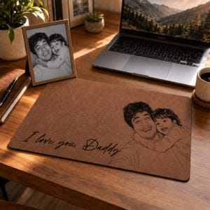 Könnte beinhalten: Eine rechteckige Schreibtischunterlage aus braunem Leder mit einer lasergravierten Illustration einer Person, die ein Kind hält. Der Text "I love you, Daddy" ist in Schreibschrift auf der linken Seite der strukturierten Unterlage eingraviert.