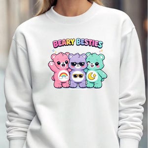May include: White sweatshirt featuring a colorful graphic of three cartoon bears with sunglasses and the text "BEARY BESTIES". The bears are pink, purple, and teal, with a rainbow, sunglasses, and a moon and star.