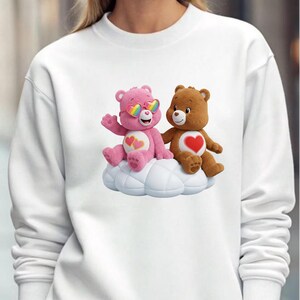 May include: White sweatshirt featuring a graphic of two Care Bears sitting on a cloud. One bear is pink and wearing rainbow heart-shaped sunglasses, the other is brown. Both bears have hearts on their chests.