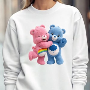 May include: White sweatshirt featuring a graphic of two Care Bears hugging. One bear is pink with a rainbow on its belly, the other is blue with a cloud and hearts design. The sweatshirt is a casual, comfortable style.