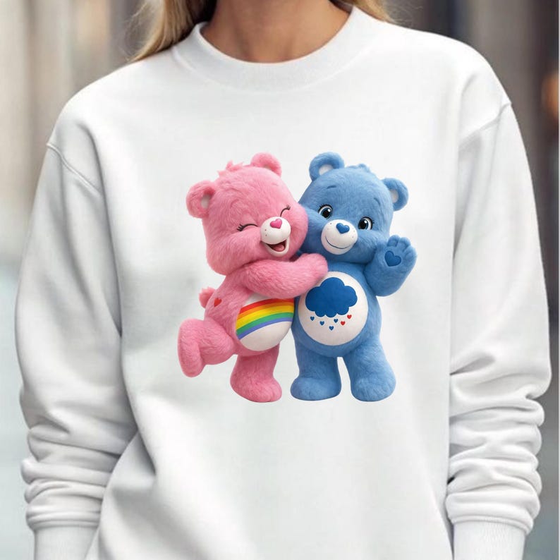 May include: White sweatshirt featuring a graphic of two Care Bears hugging. One bear is pink with a rainbow on its belly, the other is blue with a cloud and hearts design. The sweatshirt is a casual, comfortable style.