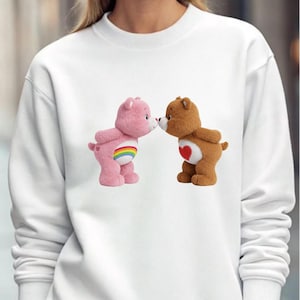 May include: White sweatshirt featuring two Care Bears. One pink bear with a rainbow belly and one brown bear with a heart on its chest are nose to nose. The sweatshirt is a solid white color.