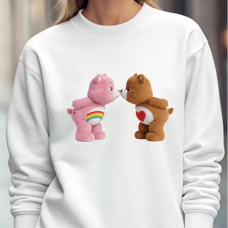 May include: White sweatshirt featuring two Care Bears. One pink bear with a rainbow belly and one brown bear with a heart on its chest are nose to nose. The sweatshirt is a solid white color.
